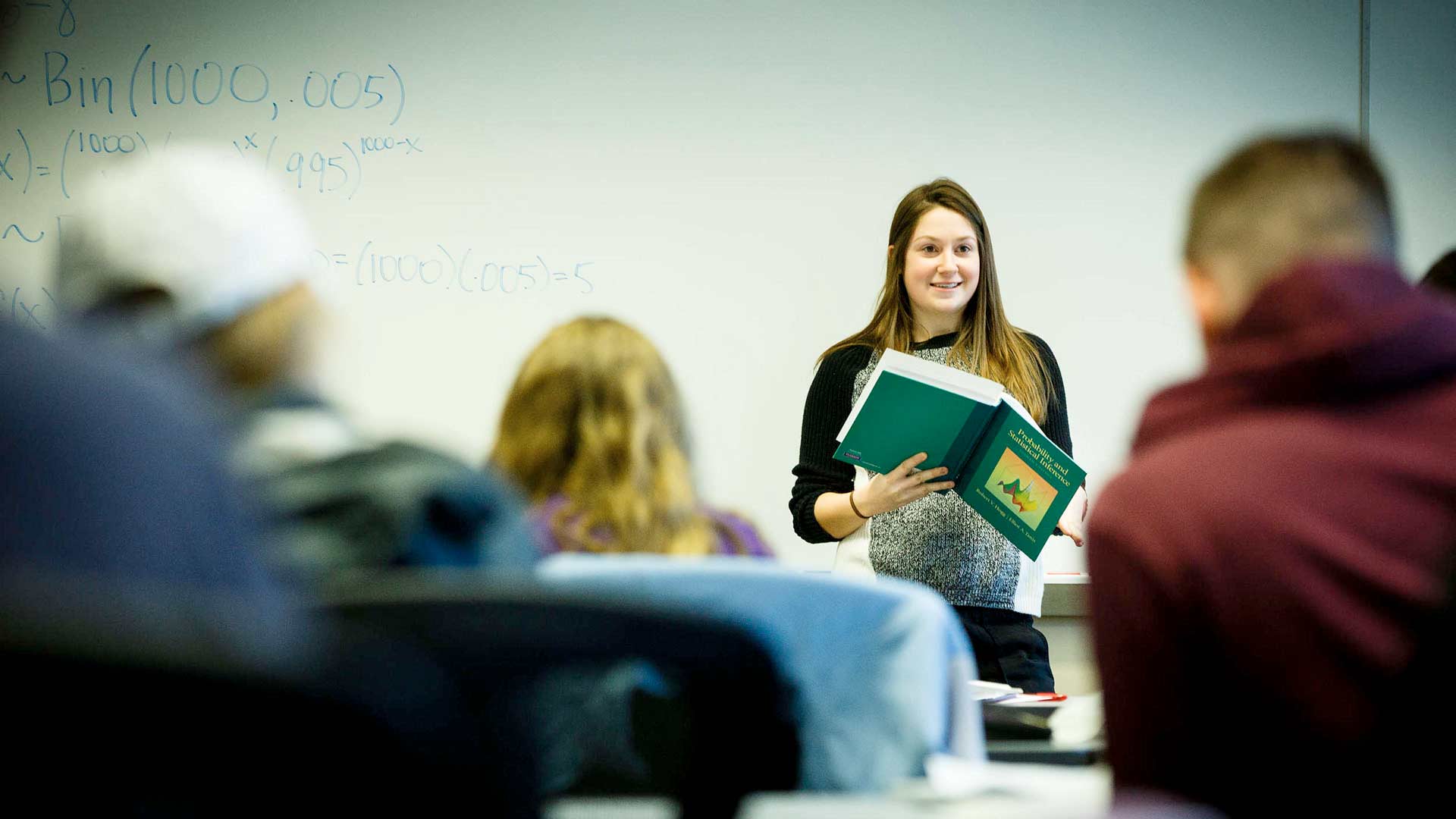 A student holds a textbook while presenting to the class during an actuarial science class.